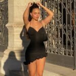Woman in black satin ruffle mini dress posing outdoors during golden hour Back view of woman in black mini dress with thigh-high boots, golden sunlight backdrop