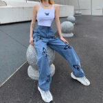 Model sitting on a concrete sphere wearing light blue wide-leg jeans with black butterfly prints and a white butterfly crop top.