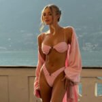 Model wearing a light pink ruffle bikini with ring detail, standing on a balcony overlooking the sea.
