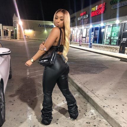 Woman wearing high waist black faux leather pants with ruched stacked leg detail, styled with crop top and heels for night out streetwear look.